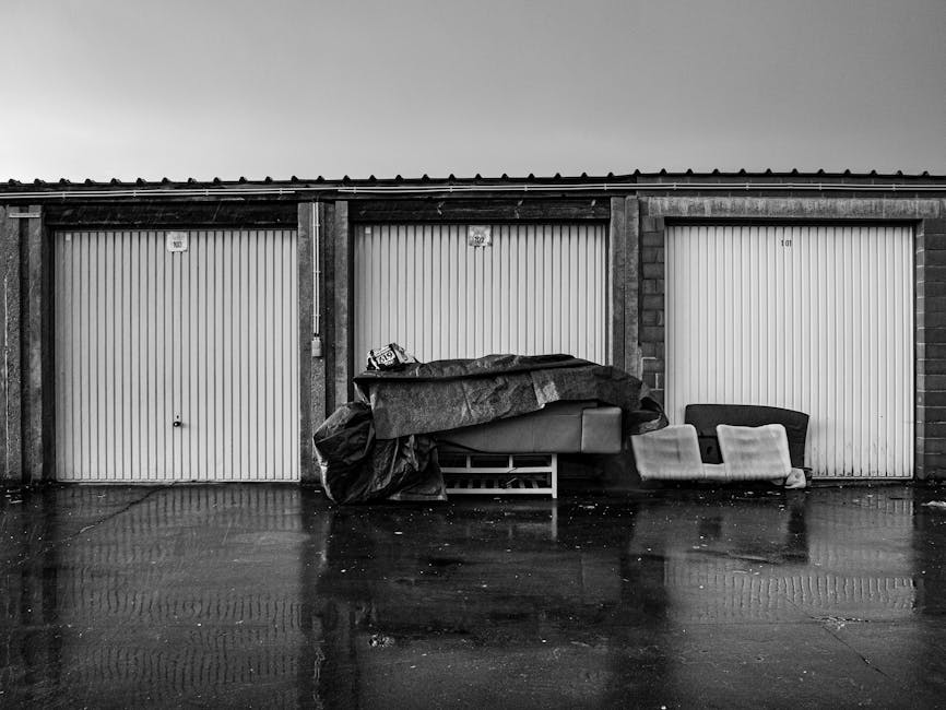 A row of three closed garage doors made of vertical metal panels with visible handles and locks, set within a brick and concrete structure. In front of the middle garage, there is a collection of discarded household items, including a large wrapped object covered with a dark tarp, partially revealing some exposed materials underneath. To the right of the wrapped item, there is a small, upholstered armchair with worn light-colored fabric and a darker wooden frame. Adjacent to the armchair, a mattress or large cushion with a light cover is leaning against the garage wall. The scene takes place outdoors on a paved surface that appears wet, creating reflections of the items and garages, with a plain, overcast sky overhead. This image relates to private rubbish collection or on-site clearance services for bulky waste, as managed by companies like Rubbish Removal Merton.
