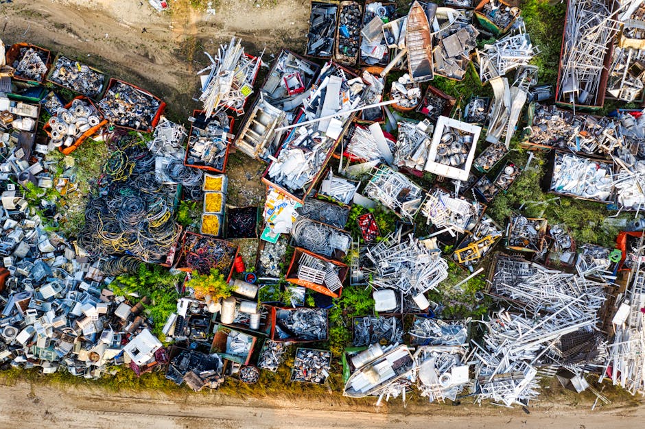 An aerial view of a large outdoor scrap metal yard featuring numerous irregularly shaped piles of assorted metallic objects. The collection includes various discarded appliances, structural metals, tubing, and wires, with some objects stacked or leaning on each other across the entire scene. The metals display diverse textures and finishes—some shiny and new-looking, others rusted and worn—ranging in colour from silver and grey to hints of brown and black. The yard is bordered by a dirt pathway visible along the bottom edge, and sparse patches of green grass and vegetation are interspersed among the scrap piles. Bright sunlight illuminates the scene, casting slight shadows that enhance the textures of the metal surfaces. This setting exemplifies an independent metal collection process, which aligns with alternative waste handling practices for metal disposal and recycling, often carried out by private rubbish removal services like Rubbish Removal Merton.