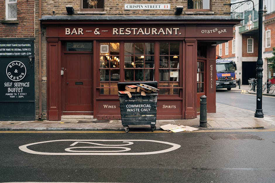 A black wheeled container with a black lid and white lettering reading 'COMMERCIAL WASTE ONLY' is positioned on the pavement in front of a storefront, partially loaded with cardboard boxes and paper debris. The container is situated directly outside a brick building with a dark red wooden facade, large multi-pane windows, and signage indicating a bar and restaurant serving wines, spirits, and cocktails. The storefront extends to the right with a narrow side door, and a small black bollard is placed nearby on the sidewalk. In the background, a street scene features a parked vehicle and bicycles, with a lamppost visible beside the building. The scene is illuminated by natural daylight, highlighting the textures of the wooden exterior and the materials in the waste container, reflecting a typical urban setting where private rubbish collection services, like those offered by Rubbish Removal Merton, may be employed for business waste disposal.