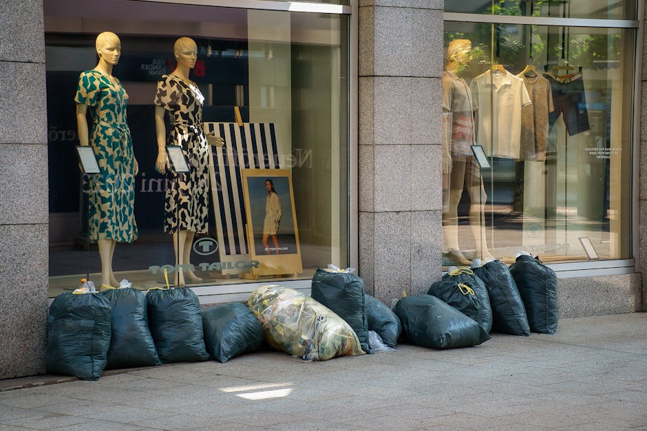 A row of black garbage bags filled with waste is positioned along the base of a large retail store window on a paved outdoor area. The bags are made of plastic and appear to be full, with some showing signs of being tightly knotted at the top. Among these bags, there is a larger, translucent bag containing mixed refuse, which is secured with clear tape. The store window behind the bags displays mannequins dressed in patterned dresses, with some mannequins having digital tablets in their hands, and clothing items such as shirts and trousers are visible on display. The window's glass reflects the surrounding urban environment and allows a view of interior fixtures and additional clothing on hangers. The stone exterior of the building features a vertical column separating the window sections. The scene is illuminated by natural daylight, creating subtle shadows on the pavement, and the setting evokes an everyday commercial streetscape where external waste disposal may be temporarily stored before collection, reflecting the activities of a commercial waste management service like Rubbish Removal Merton.