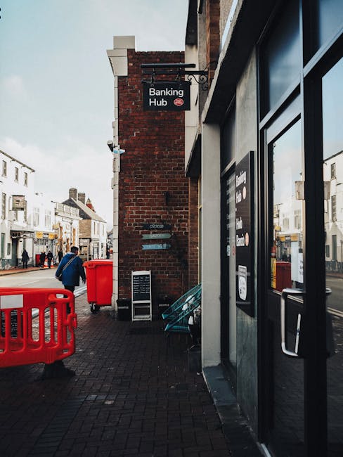 A black wheeled container with a black lid and white lettering reading 'COMMERCIAL WASTE ONLY' is positioned on the pavement in front of a storefront, partially loaded with cardboard boxes and paper debris. The container is situated directly outside a brick building with a dark red wooden facade, large multi-pane windows, and signage indicating a bar and restaurant serving wines, spirits, and cocktails. The storefront extends to the right with a narrow side door, and a small black bollard is placed nearby on the sidewalk. In the background, a street scene features a parked vehicle and bicycles, with a lamppost visible beside the building. The scene is illuminated by natural daylight, highlighting the textures of the wooden exterior and the materials in the waste container, reflecting a typical urban setting where private rubbish collection services, like those offered by Rubbish Removal Merton, may be employed for business waste disposal.