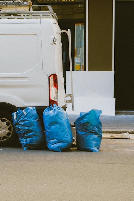Photograph showing the rear side of a white commercial van parked on a city street with three large blue rubbish bags placed at the back near the vehicle's rear door. The bags are made of thick plastic with a slightly crinkled texture, and vary in shades of blue, with one having a darker hue. The background features a building with a glass window and a white structural element, possibly a step or platform, positioned on the sidewalk. The scene appears to capture an instance of private waste disposal possibly handled by a rubbish removal service such as Rubbish Removal Merton, emphasizing independent collection of waste materials outside a commercial or residential property. The street surface is asphalt, and the lighting suggests a clear day with natural light illuminating the scene. The arrangement indicates an intention to load or unload waste, consistent with rubbish collection activities often performed by local waste removal companies.