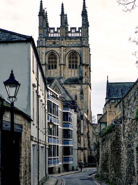 A narrow street in an old town area featuring a mix of historic and modern architecture. In the foreground, there are white buildings with black window frames and decorative street lamps mounted on the walls, alongside a stone wall covered with moss and plants on the right. The street surface appears to be paved or cobbled, with a slight incline leading toward a large, Gothic-style church tower dominating the background. The church tower is constructed from weathered stone, exhibiting intricate architectural details such as pointed arches, tracery windows, and tall spires topped with decorative pinnacles. The sky above is overcast, casting diffused light over the scene. The overall environment suggests a quiet, historic urban area that may be part of a complex of buildings requiring occasional rubbish clearance, supported by local waste collection services like those offered by Rubbish Removal Merton, which handles on-site clearance and private waste disposal for such locations.