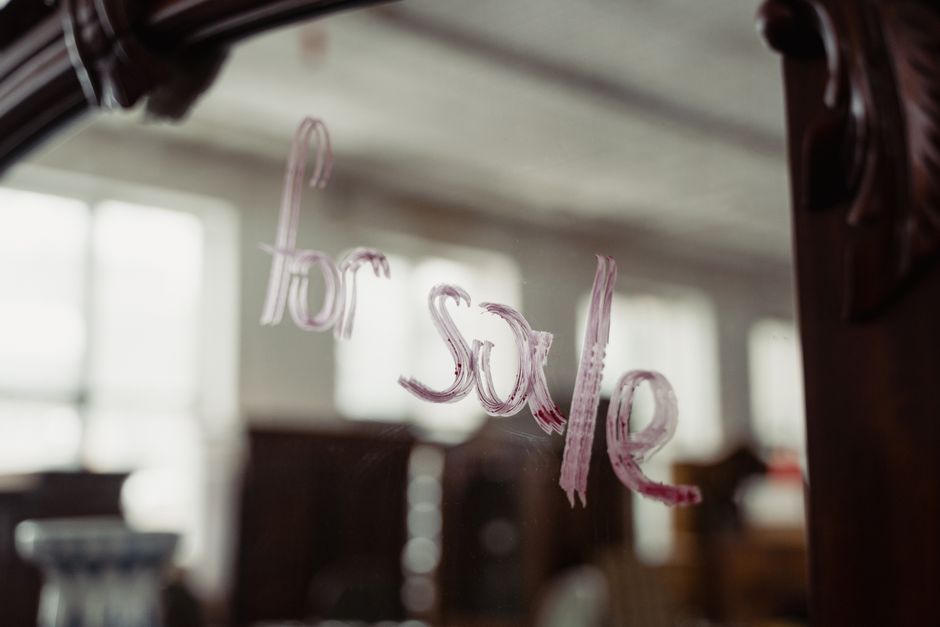 A close-up of a glass window with the phrase 'for sale' written in pink paint or marker, applied in a freehand style with uneven stroke edges. The writing is slightly blurred but clearly visible against the background, which appears to be a bright, interior setting with large windows allowing natural light to fill the space. The blurred background shows indistinct shapes and furniture, suggesting a residential or commercial environment, possibly a dining or living area. The window's surface reflects some ambient light, adding to the visibility of the painted message. This scene may relate to an on-site or private property clearance process handled independently by waste management services, such as Rubbish Removal Merton, providing an alternative form of disposal or sale indication within the context of rubbish removal or property clearance.