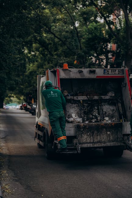 A refuse collection worker wearing a green uniform and protective helmet is standing on the rear platform of a white waste collection van, which is stationary on a residential street. The open back of the vehicle reveals a large, blackened and slightly rusty container used for collecting rubbish, with visible dirt and debris inside. The van is parked close to the curb, with a background of leafy trees providing partial shade over the scene and other vehicles parked along the road. The worker appears to be in the process of emptying or inspecting the waste compartment, indicating an on-site waste collection or rubbish removal activity. Surrounding environment includes a sidewalk with foliage and a quiet neighborhood setting, reflecting typical urban rubbish management operations that may involve independent collection services like those offered by Rubbish Removal Merton, in contrast to council-led disposal methods.