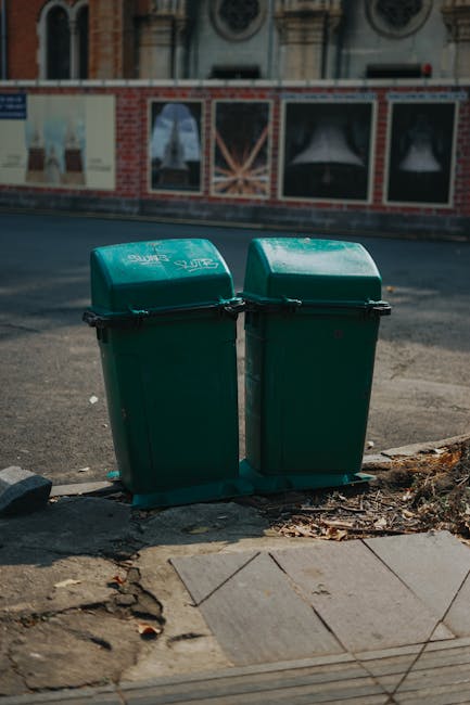 Two identical green wheeled rubbish bins with hinged lids are positioned side by side on a paved sidewalk at the edge of a street. The bins are made of durable plastic with a slightly textured finish and are branded with white text and graphics, indicating they are for waste disposal. The bases of the bins rest on a black rubber mat to keep them stable. Behind the bins, a brick wall features a series of large posters or advertisements framed by metal borders, with some windows and an upper building façade visible in the background. The street surface appears to be asphalt, with a small patch of uneven pavement and some loose debris including fallen leaves and small stones surrounding the bins. Natural daylight illuminates the scene, casting soft shadows and highlighting the clean, functional appearance of the waste containers, which are part of local waste management provisions, possibly set out for collection or independent disposal services, reflecting the typical urban environment in Merton.