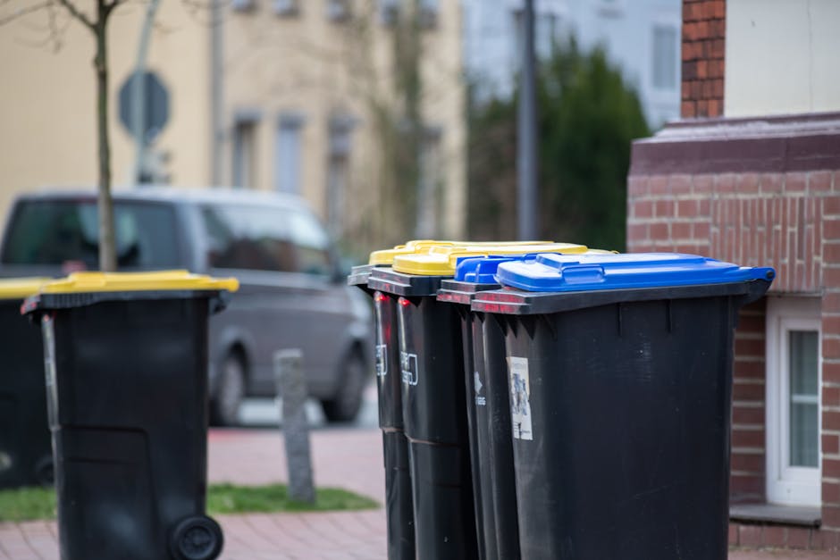 In the foreground, four large black wheeled rubbish bins with textured plastic surfaces are lined up on a paved sidewalk next to a red-brick building. Each bin has a contrasting coloured lid: two yellow, one blue, and one dark blue, all with smooth finishes and slightly raised edges. The bins are positioned close together, with the yellow-lidded bin slightly to the left and the blue-lidded bin toward the center. Behind them, a parked dark grey van with a shiny, metallic finish is partially visible, out of focus. The background features a residential street scene with a few trees, a lamppost, and more houses with neutral-colored walls and windows, suggesting an urban or suburban area. The overall lighting indicates daytime with natural light illuminating the scene evenly. This image likely depicts the typical arrangement of waste collection containers on a residential street, associated with waste management and rubbish removal services that operate independently of local authority collection, as represented by Rubbish Removal Merton.
