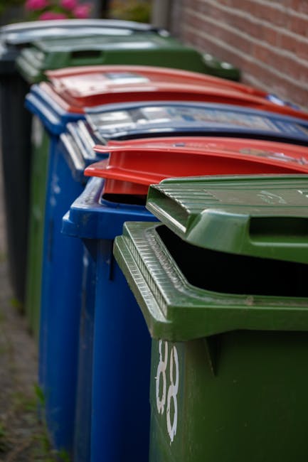 Two identical green wheeled rubbish bins with hinged lids are positioned side by side on a paved sidewalk at the edge of a street. The bins are made of durable plastic with a slightly textured finish and are branded with white text and graphics, indicating they are for waste disposal. The bases of the bins rest on a black rubber mat to keep them stable. Behind the bins, a brick wall features a series of large posters or advertisements framed by metal borders, with some windows and an upper building façade visible in the background. The street surface appears to be asphalt, with a small patch of uneven pavement and some loose debris including fallen leaves and small stones surrounding the bins. Natural daylight illuminates the scene, casting soft shadows and highlighting the clean, functional appearance of the waste containers, which are part of local waste management provisions, possibly set out for collection or independent disposal services, reflecting the typical urban environment in Merton.