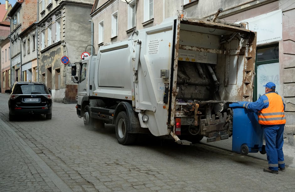 A white waste collection truck parked on a narrow urban street with cobblestone pavement, operated by Rubbish Removal Merton. The truck's rear is open, revealing mechanical grinding and compacting mechanisms for in-situ rubbish processing. A worker dressed in blue and orange safety clothing, including gloves and a cap, is seen depositing waste into a blue wheeled bin positioned next to the truck. The street is lined with aged, multi-story terraced buildings with stone facades and large windows, some featuring old paintwork and minor weathering. A black vehicle is parked further down the street, and traffic signs, including a no-parking sign, are mounted on the buildings. The scene is lit with natural daylight, creating clear visibility of the waste collection operation in a typical residential area, highlighting the process of private or independent rubbish removal in an urban setting.