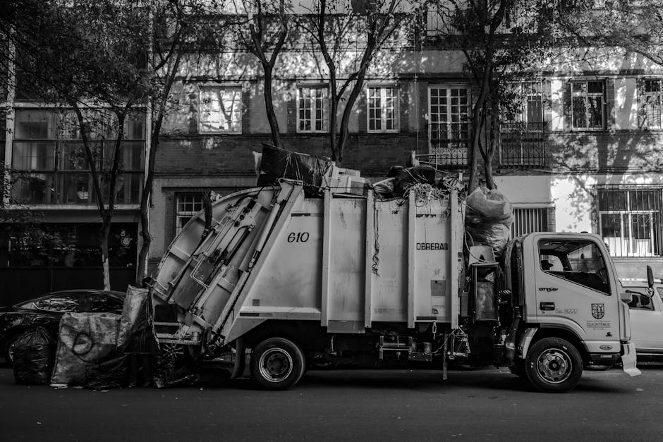 A side view of a large commercial rubbish collection truck parked on a residential street. The truck has a white body with some visible signs of wear and is labeled with the number 610 and the word 'OBREAS' on its side. The loading bay at the rear is filled with an assortment of waste materials, including black plastic garbage bags, cardboard boxes, and miscellaneous debris. The truck is positioned on a paved road, with a row of multi-story residential buildings featuring brick facades, large windows, and balconies visible in the background. Several leafless trees cast shadows across the buildings and the parked truck, suggesting a late autumn or winter setting. The scene is captured in black and white, emphasizing the textures of the waste, the truck's metal surfaces, and the surrounding environment, illustrating an example of private waste collection as part of rubbish removal services provided by Rubbish Removal Merton.