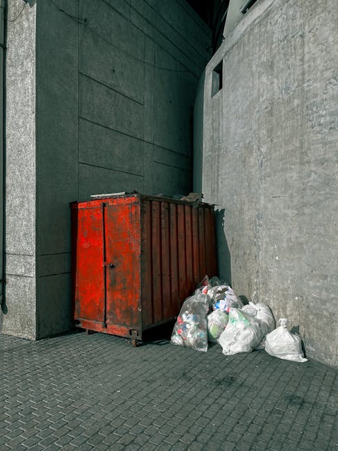 In an urban outdoor setting, a large, rectangular, metal rubbish skip with a weathered red finish and visible rust spots is positioned against a dark, smooth wall on the left and a rough, light grey concrete wall on the right. The skip's surface appears textured with peeling paint, indicating extensive use. Surrounding the base of the skip are several loosely tied plastic rubbish bags filled with assorted waste materials, including paper, plastic bottles, and other household refuse. The bags are semi-transparent and lie on a dark, textured paving surface that extends into the background. The environment is minimally lit, with natural light highlighting the subtle variations in the wall surfaces and the details of the bags and skip. The scene suggests a commercial or private property area where independent rubbish collection or on-site waste management services, such as those provided by Rubbish Removal Merton, are used for waste disposal outside standard municipal services.