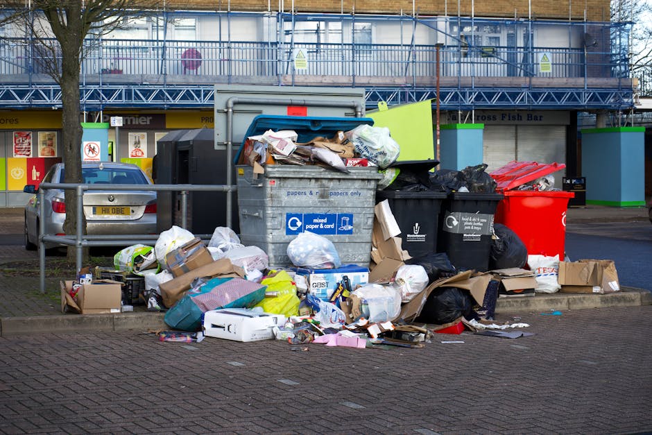 A collection of overflowing rubbish bins and surrounding waste materials situated on a paved area next to a parking space. There are multiple black, red, and gray bins, with some lids open, revealing waste such as paper, cardboard boxes, plastic bags, and assorted packaging. Additional loose items, including flattened cardboard boxes, plastic containers, and discarded paper, are scattered on the ground around the bins. In the background, a commercial building with a blue metal framework and a yellow storefront features windows and signage, while a parked silver car with a yellow license plate is visible behind a short metal railing. The scene is outdoors under daylight, with minimal environmental context beyond the immediate waste area. The setting appears to be a public space possibly serviced by private waste removal, aligning with the themes of alternative rubbish collection or on-site clearance often handled by companies like Rubbish Removal Merton.