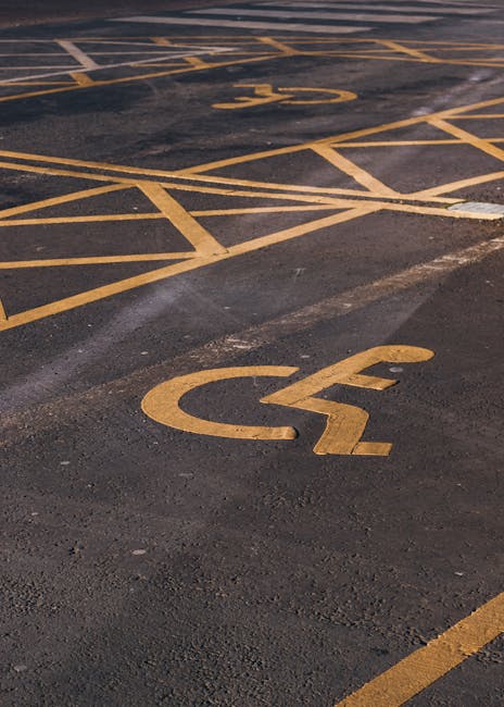 A photograph of a parking lot surface displaying bright yellow painted markings for disabled parking spaces and accessibility symbols. The foreground shows a prominent wheelchair symbol painted directly onto the dark asphalt, with clear, solid lines forming the wheelchair shape, including the circular wheel and platform. In the background, additional yellow lines delineate individual parking bays, some of which are designated for disabled parking, indicated by the wheelchair symbols and striped markings. The asphalt surface appears slightly textured, with areas of wear and minor discoloration, suggesting regular use. The lighting in the scene is even, highlighting the contrast between the dark asphalt and the vivid yellow lines, making the markings easily visible. This setting exemplifies on-site parking area markings that support alternative waste handling logistics by ensuring accessible vehicle parking, an aspect that local waste management companies like Rubbish Removal Merton might consider when planning waste collection routes from accessible locations, especially in premises providing private or independent waste disposal arrangements.
