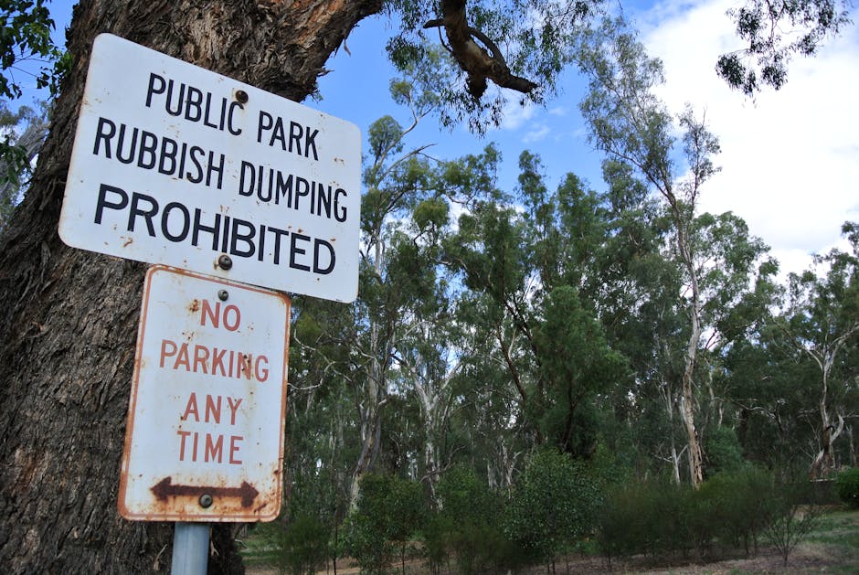 A close-up view of two weathered metal signs attached to a tree trunk in an outdoor park setting. The upper sign, predominantly white with black text, reads 'PUBLIC PARK RUBBISH DUMPING PROHIBITED' and is slightly rusty with some minor chipping around the edges. The lower sign, white with faded red lettering, states 'NO PARKING ANY TIME' and features a left-pointing arrow. The signs are positioned on the left side of the image, with the tree's rough bark framing them. In the background, a dense cluster of tall, slender eucalyptus trees with light green foliage extends upward, contrasting with a partly cloudy sky. The environment suggests an area where visitors may be discouraged from improper waste disposal or parking in violation of rules, emphasizing the importance of responsible rubbish removal and site management. Rubbish Removal Merton offers professional services aligned with these practices, supporting private waste collection and on-site clearance needs in such natural or community spaces.