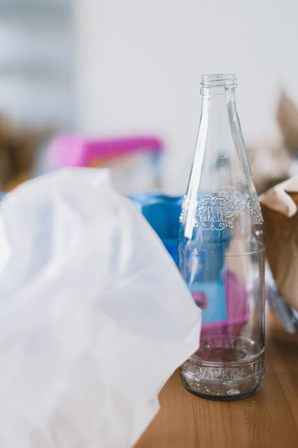 A clear glass soda bottle with embossed decorative pattern and rounded shoulders, positioned upright on a wooden surface. The bottle is empty or contains minimal residual liquid and is surrounded by various discarded packaging materials, including a crumpled white plastic bag in the foreground and other partially visible items such as a blue container and brown paper packaging towards the background. The setting appears to be indoors, likely in a domestic environment, with light-colored walls and soft, natural lighting illuminating the scene. The scene reflects typical waste that may be collected during an on-site rubbish clearance or private waste handling service provided by Rubbish Removal Merton, fitting within the category of general rubbish removal in a residential context.