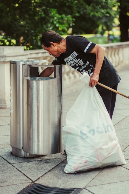 A middle-aged man wearing a black T-shirt with white text, black trousers, and while holding a wooden broom, is seen disposing of a large white trash bag filled with waste into a stainless steel public rubbish bin. The bin has a sleek, cylindrical design with a polished finish and a rectangular opening at the top for waste insertion. It is situated on a paved surface, likely a sidewalk or a park pathway, with a sturdy stone or concrete balustrade running alongside. In the background, there are lush green trees and a well-maintained grassy area, suggesting an outdoor setting in a park or public space. The lighting appears natural, indicating daytime with ambient light illuminating the scene. The overall scene reflects independent waste disposal or on-site rubbish management, which Rubbish Removal Merton might support through alternative collection services in a community environment.