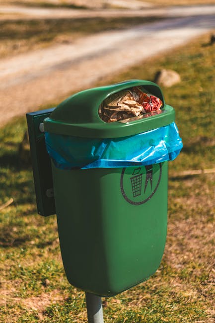 A small, green, outdoor rubbish bin mounted on a metal post, with a semi-circular, lid featuring a transparent window showing mixed waste including plastic wrappers and paper inside. The bin has a blue plastic liner partially visible beneath the lid and a black icon printed on the front, indicating waste disposal. It is situated on a grassy area with a dirt pathway and blurred natural background, suggesting a park environment. The scene is illuminated by natural sunlight, highlighting the textured surface of the bin and the surroundings. The image reflects the importance of proper waste disposal and is relevant to waste management services such as those offered by Rubbish Removal Merton, emphasizing the need for responsible rubbish collection and disposal outside of traditional municipal channels.