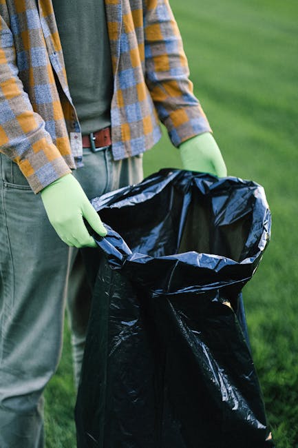 A person wearing a checkered shirt with yellow, blue, and gray patterns, along with a green long-sleeve top underneath, is standing outdoors on a grassy area. They are holding open a large black plastic rubbish bag, which is partially crumpled, with both hands covered in light green gloves. The background features lush green grass, indicating an outdoor setting. The scene suggests waste collection or rubbish disposal, consistent with services offered by Rubbish Removal Merton, highlighting the process of segregating or collecting waste for disposal or recycling, possibly as part of an on-site clearance or private waste handling activity.