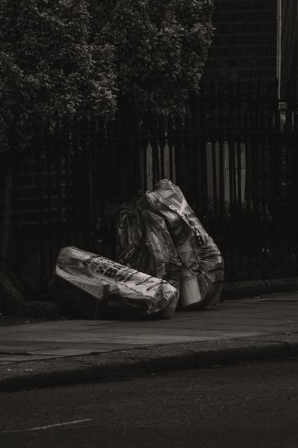 Two large black refuse bags filled with waste are placed on the edge of a sidewalk in front of a wooden fence with vertical slats, surrounded by trees and foliage. The bags appear to be made of thick plastic and are crumpled with visible wrinkles and folds. They are positioned on a paved surface, likely a street or footpath, with part of an asphalt road visible in the foreground. The scene is outdoors, with natural light casting gentle shadows, indicating daytime. The setting suggests a local rubbish removal scenario, typical for private or independent waste collection services, aligning with the context of alternative waste disposal methods managed by companies like Rubbish Removal Merton.